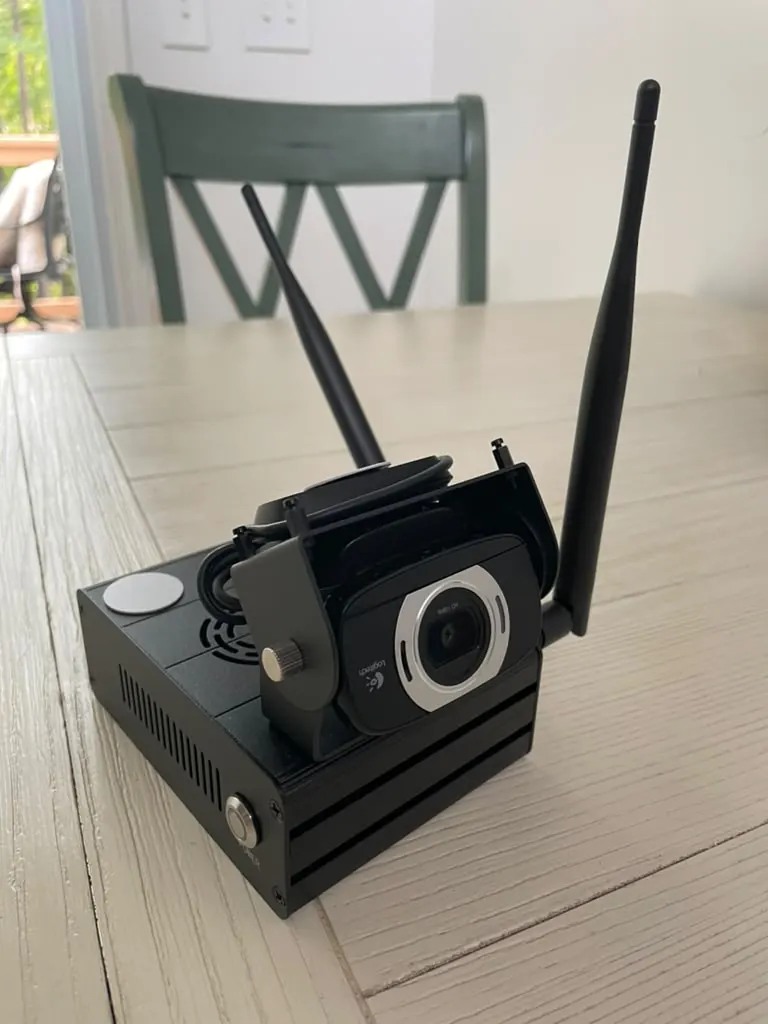 The Pasiero home hub: a compact black metal box with two Wi-Fi antennas, a round tap pad on top, and a clip-on camera.